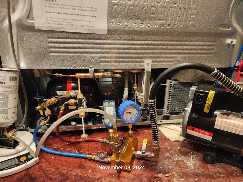 Refrigerator compressor with gauges and tools in a repair setup. Visible wiring, pressure gauges, and tubing indicate maintenance work. Note with technical instructions and caution warnings attached.