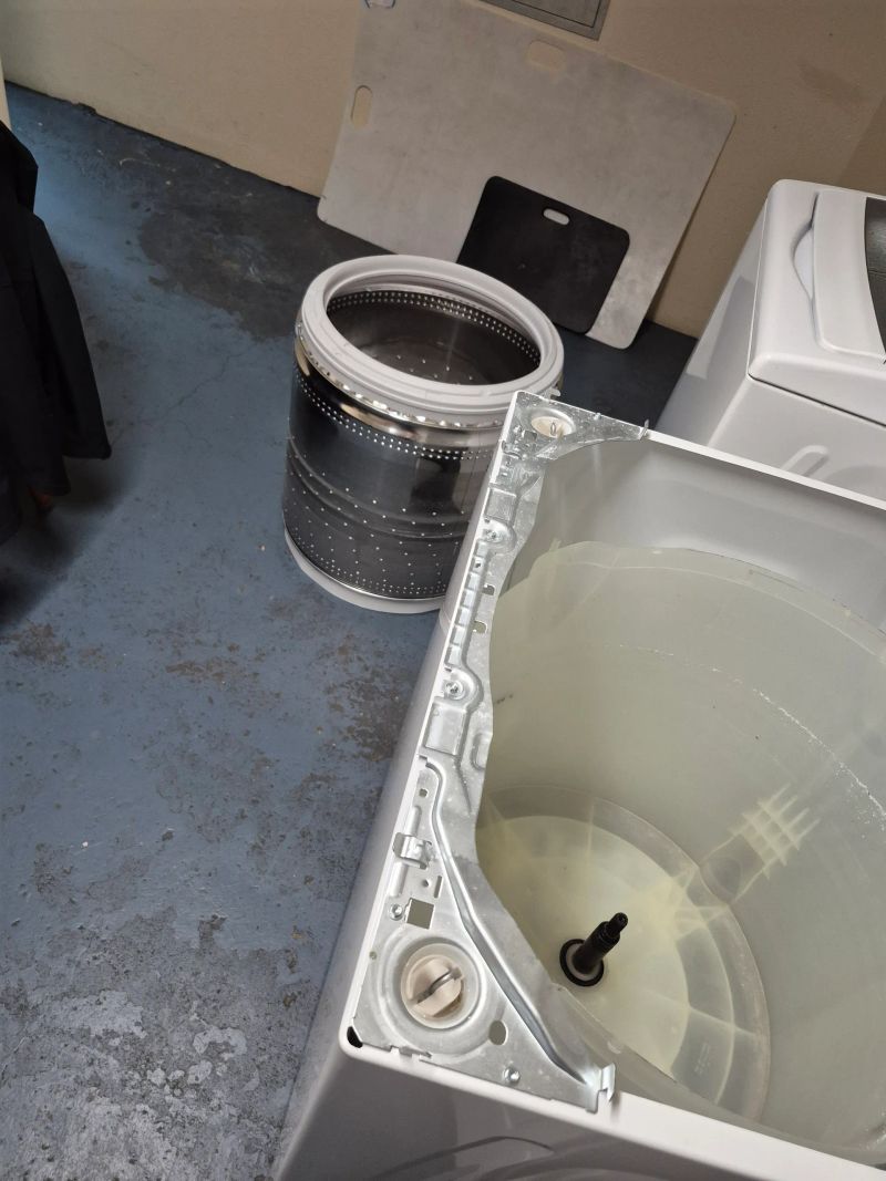 The interior of a washing machine with its drum removed, showing the central spindle and internal components, placed on a concrete floor with other laundry appliances nearby.