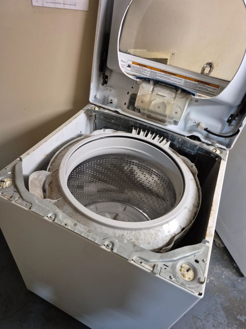 Open top-loading washing machine with visible drum and detached top lid, showing interior and some dirt or dust inside.