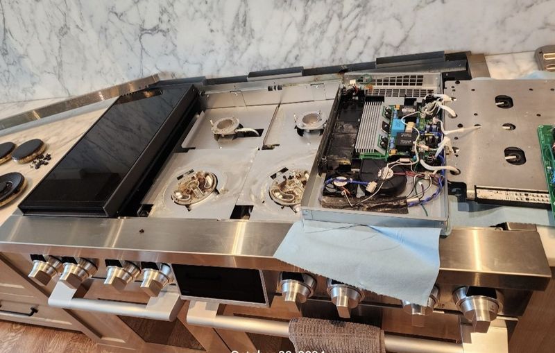 A partially disassembled stainless steel kitchen stove with exposed wiring and components, placed on a marble countertop.